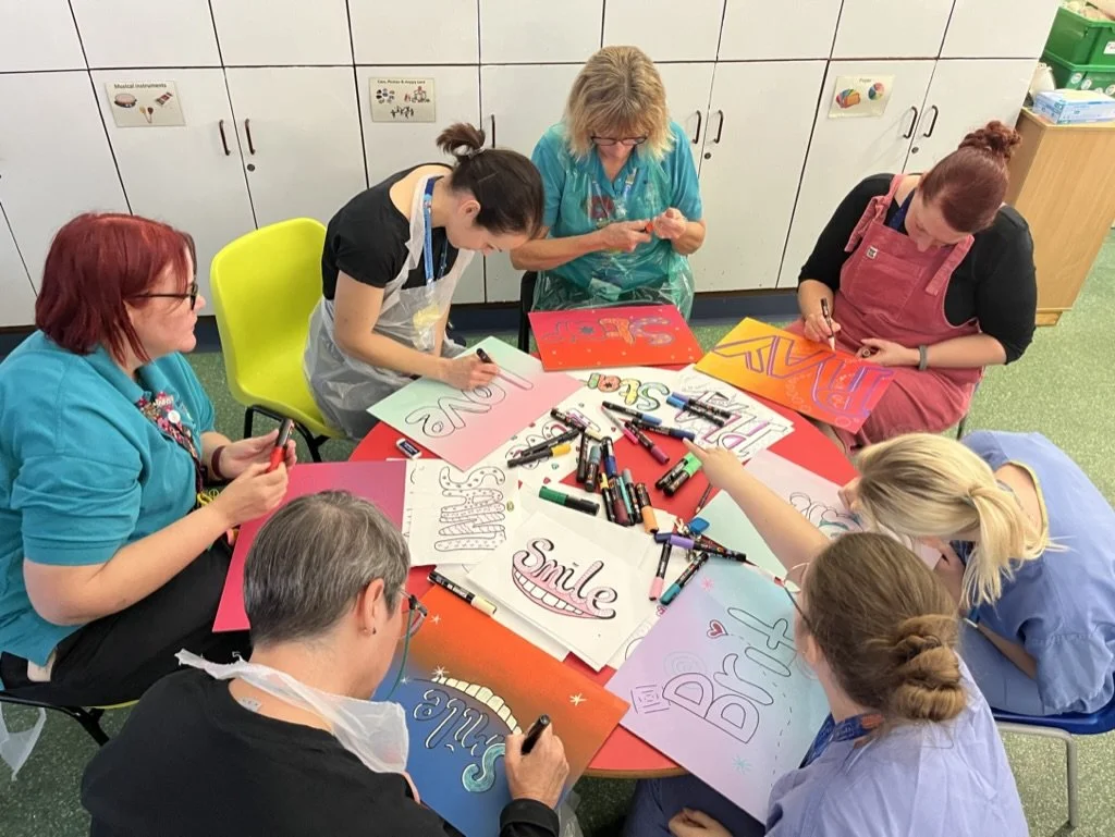 Group of people sitting around a red table making colorful posters with markers, some posters read 'Smile,' 'Dream,' 'Love,' 'Peace,' and 'Diversity' in a classroom or craft room.