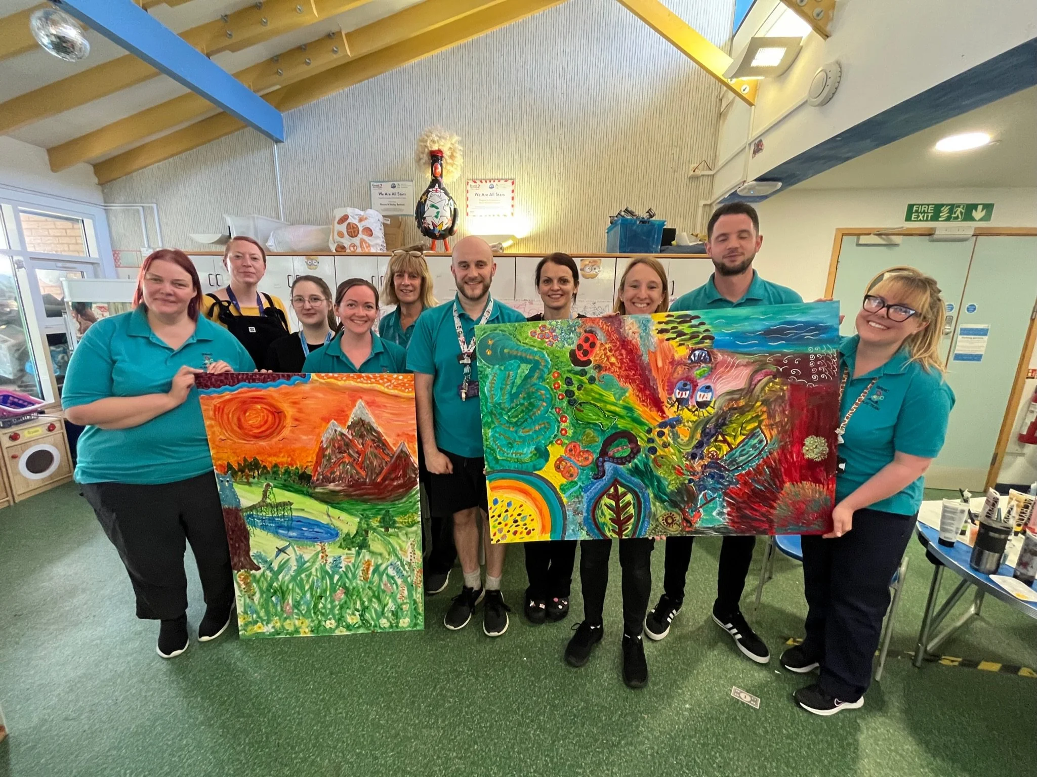 Group of eight people holding colorful, abstract paintings in an indoor space with yellow and blue decor, cabinets, and a fire exit sign.