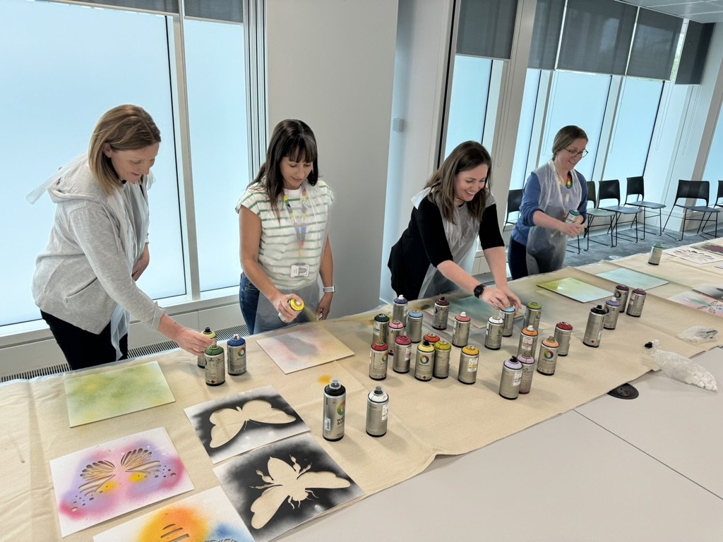 Four women spray painting artwork on paper at a table in a bright room with large windows.