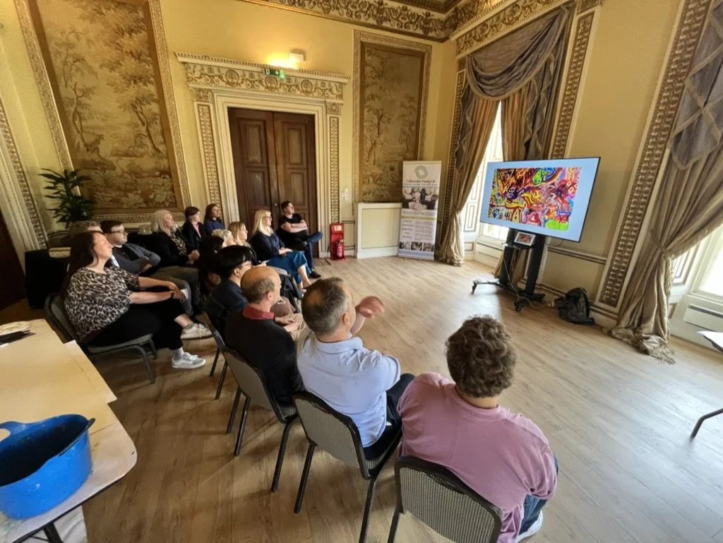 A group of people sitting on chairs in a decorated room watching a colorful digital presentation on a large screen.