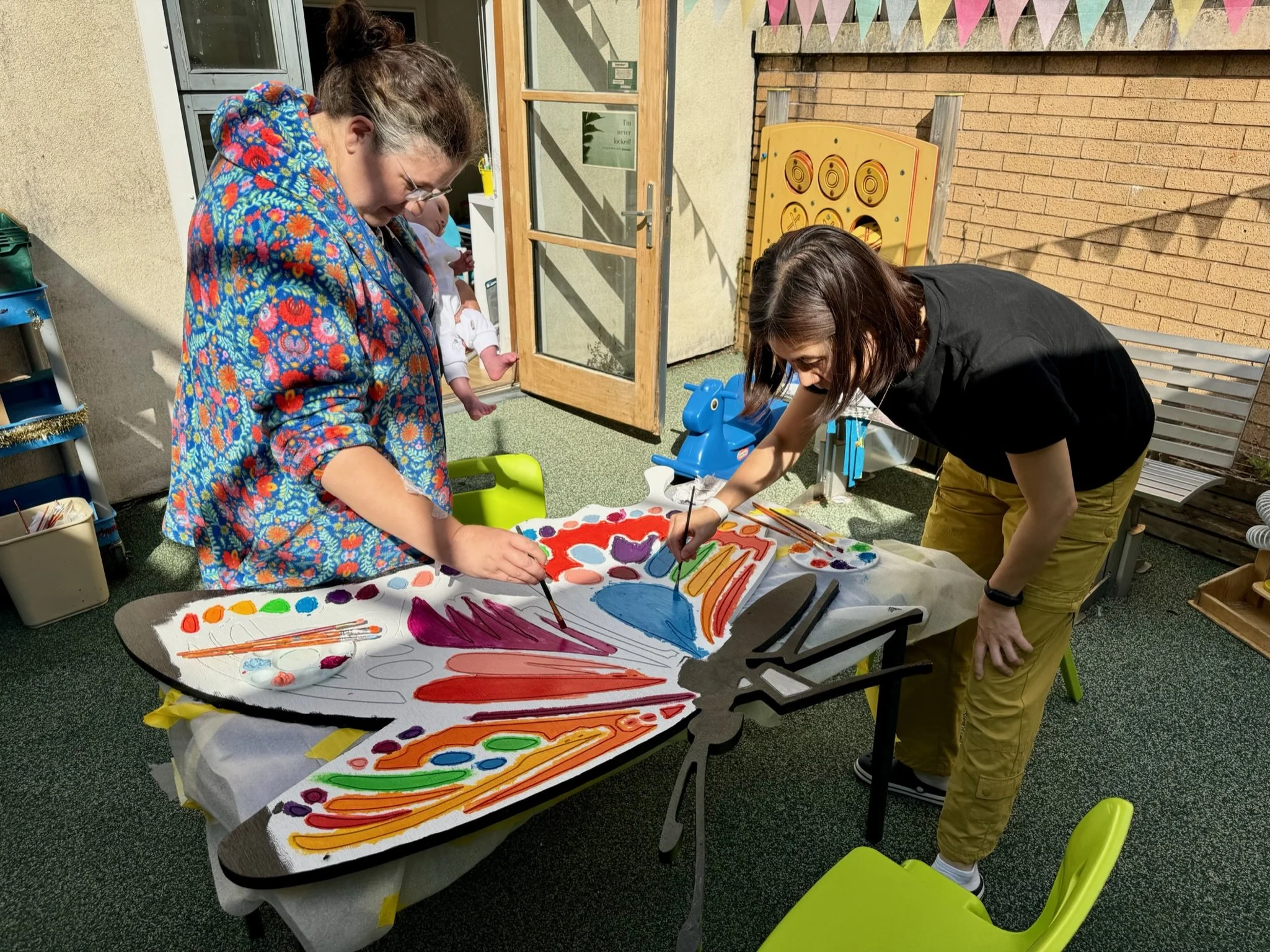 Two women are painting a colorful artwork on a large sheet of paper at a table outdoors. One woman has dark hair and glasses, wearing a floral hoodie, and the other has dark hair and is dressed in black and yellow. There are painting supplies and a b