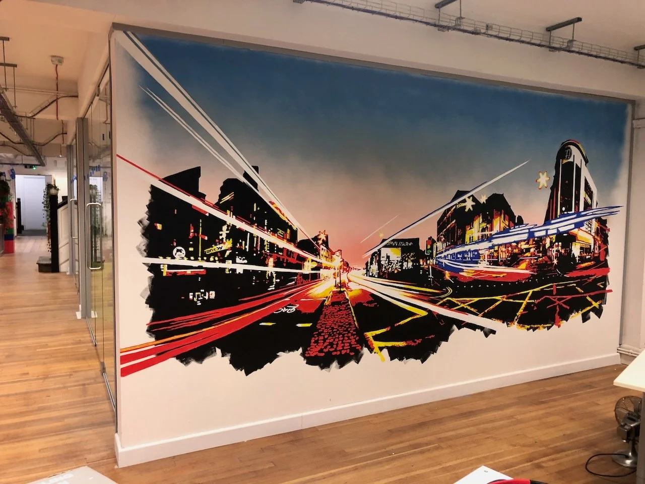 Cityscape mural of a vibrant urban street scene at dusk with light streaks, buildings, and a sunset sky, painted on an interior wall.