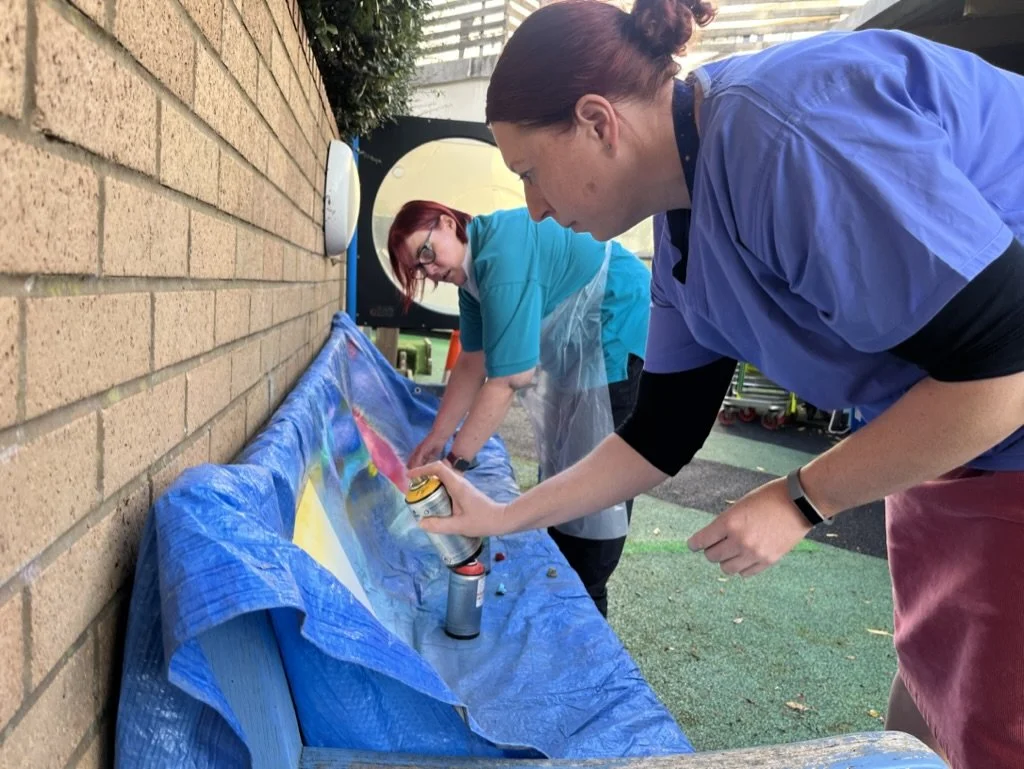 Two women spray paint a large surface placed on a blue tarp against a brick wall outdoors.
