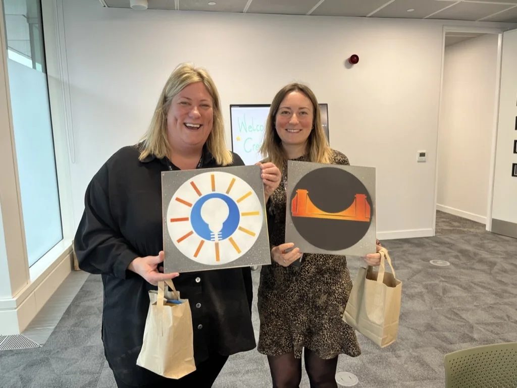 Two women smiling and holding up square signs with graphic designs inside a modern office space.