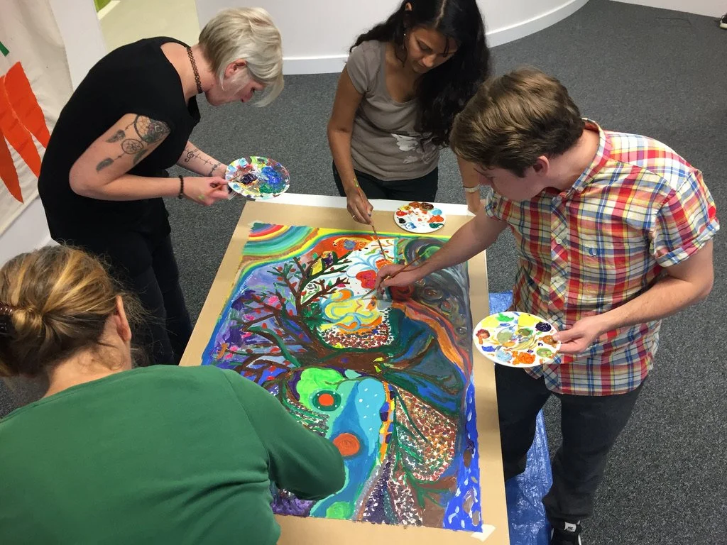 Four people are gathered around a table, working on a colorful, abstract painting of a tree with rainbow-colored sky and swirling patterns.