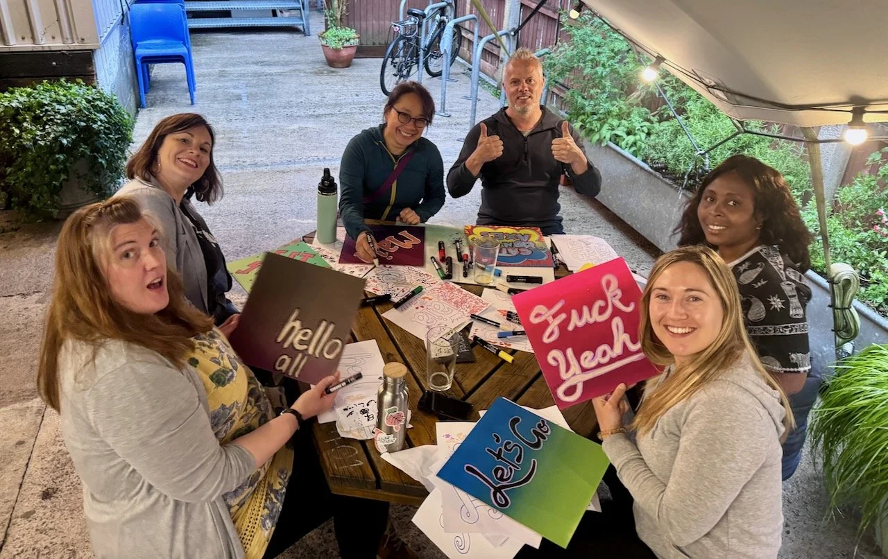 Group of six women sitting around a table outdoors, enjoying a creative night with markers, posters, and signs that read 'hello all,' 'Let's care,' and 'Fuck Yeah.' Some women are smiling and holding signs, and one man at the back gives a thumbs-up.