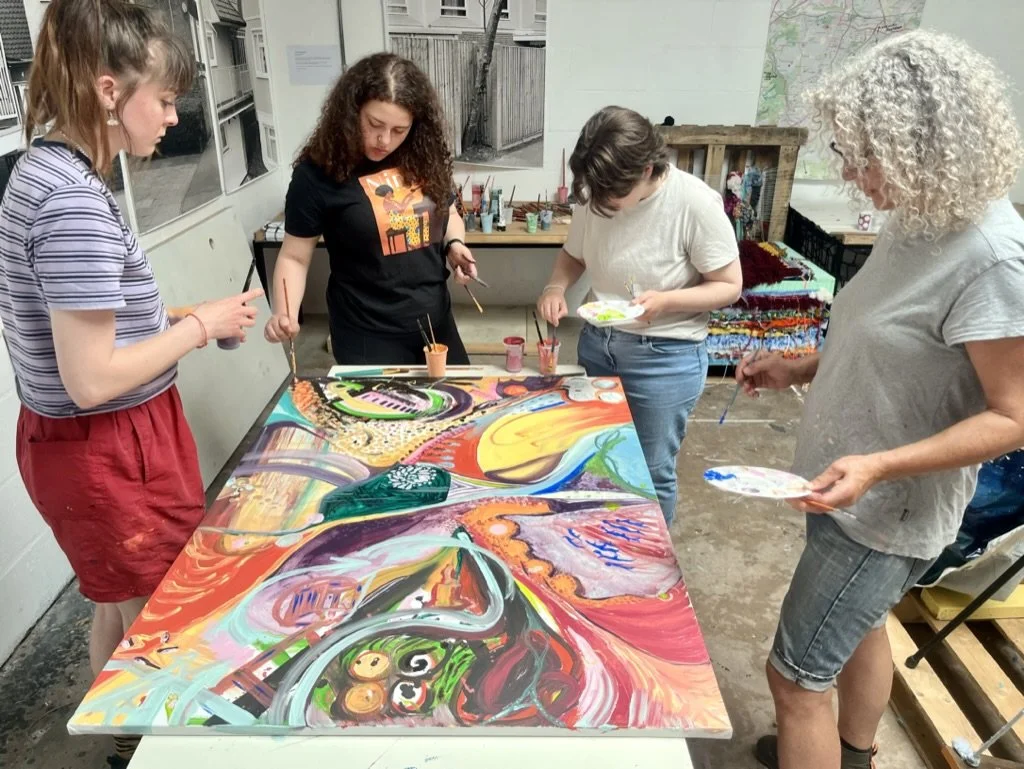 Four women and girls are painting an abstract, colorful artwork on a large canvas in an art studio.