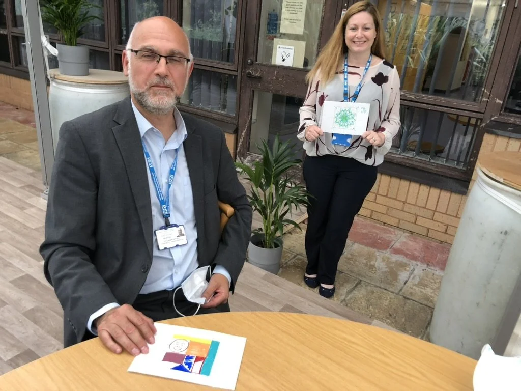 A man with glasses and a beard in a suit sits at a table holding a face mask, with drawings on paper in front of him. A woman with long blonde hair stands behind him, smiling, holding a colorful mandala-like artwork. They are outside, with potted pla