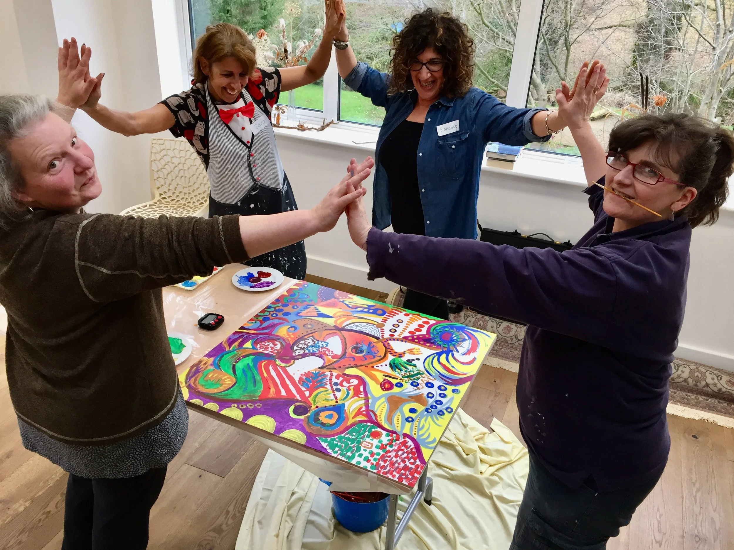 Four women celebrating around a colorful abstract painting during an art event in a bright room.