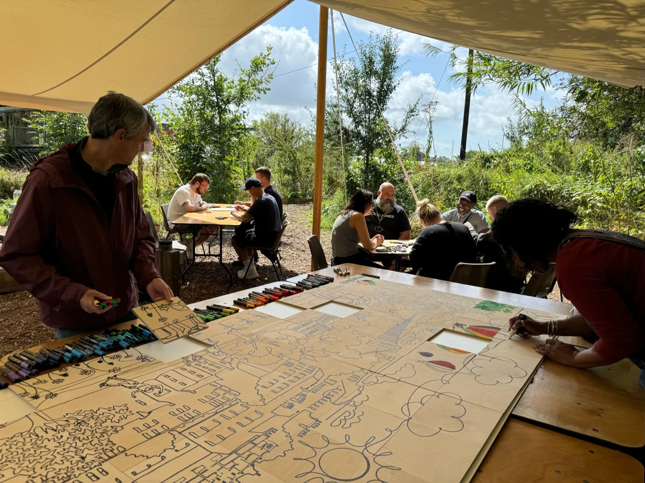 People sitting and drawing on a large paper under a tent in an outdoor setting.