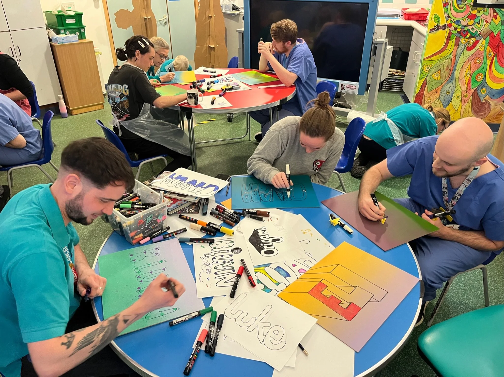 People sitting around a table creating colorful posters with markers, with art supplies scattered on the table, in a room decorated with vibrant artwork.