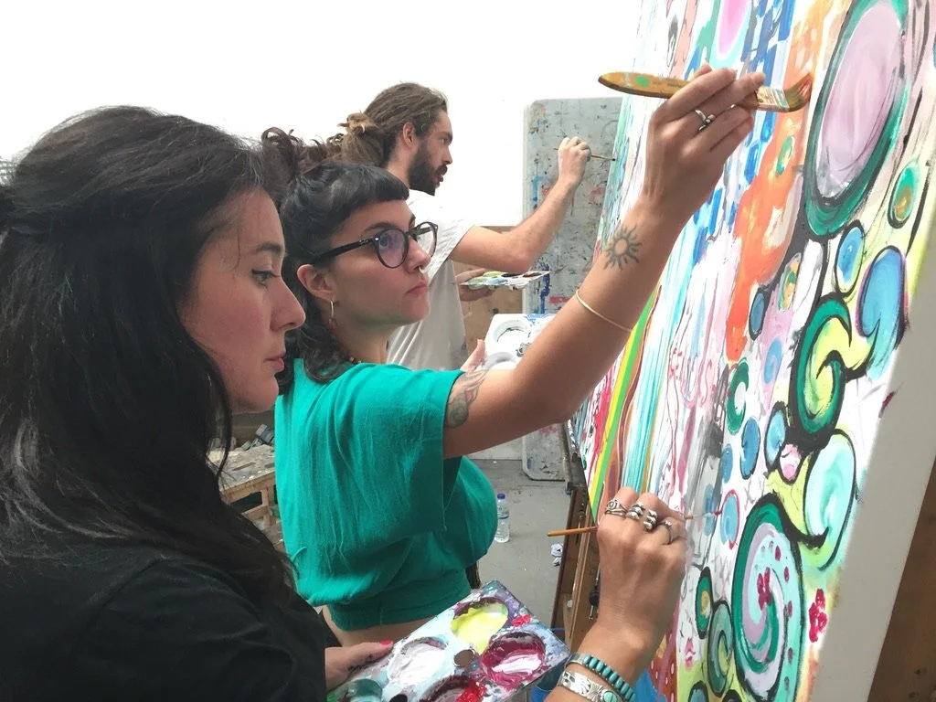 Three people painting a colorful abstract mural on a large canvas, focused on their work.