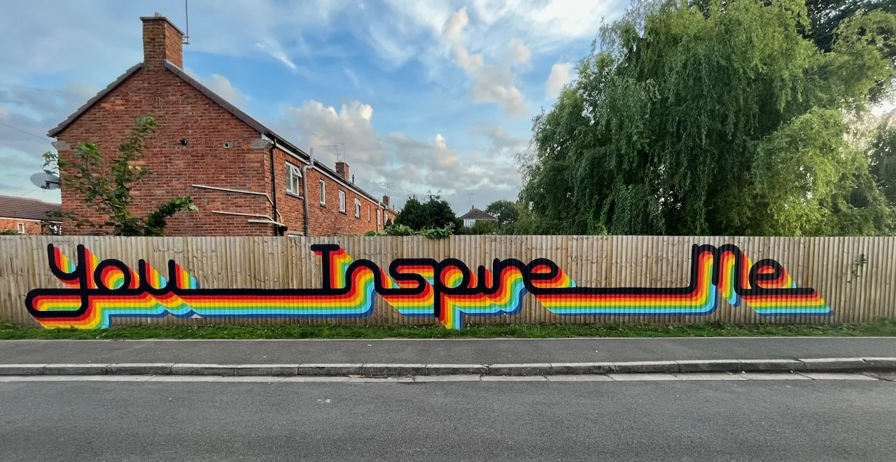 Colorful rainbow-style graffiti on wooden fence reads 'You Inspire Me' with trees, house, and cloudy sky in background.