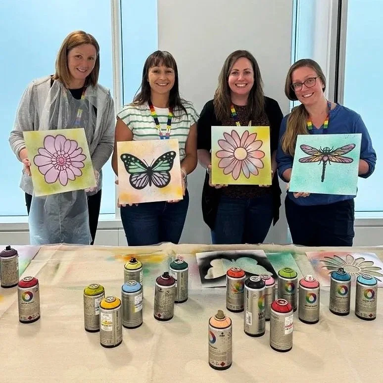 Four women standing behind a table, each holding art prints of a flower, butterfly, flower, and dragonfly. Paint cans and art supplies are on the table in front of them.