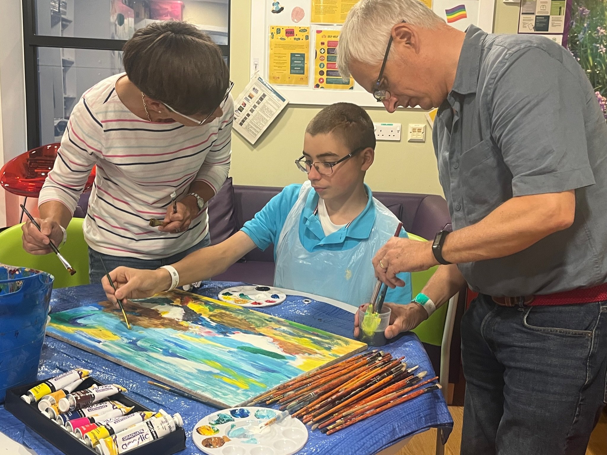 Two adults and a young man are painting together at a table covered with colors and brushes in an art classroom or therapy room.