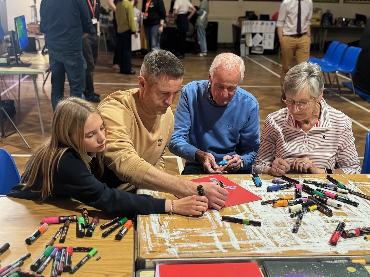 Four people, including a young girl, middle-aged man, elderly man, and elderly woman, are seated at a table drawing with markers on red paper in a busy indoor setting, with other people and chairs visible in the background.