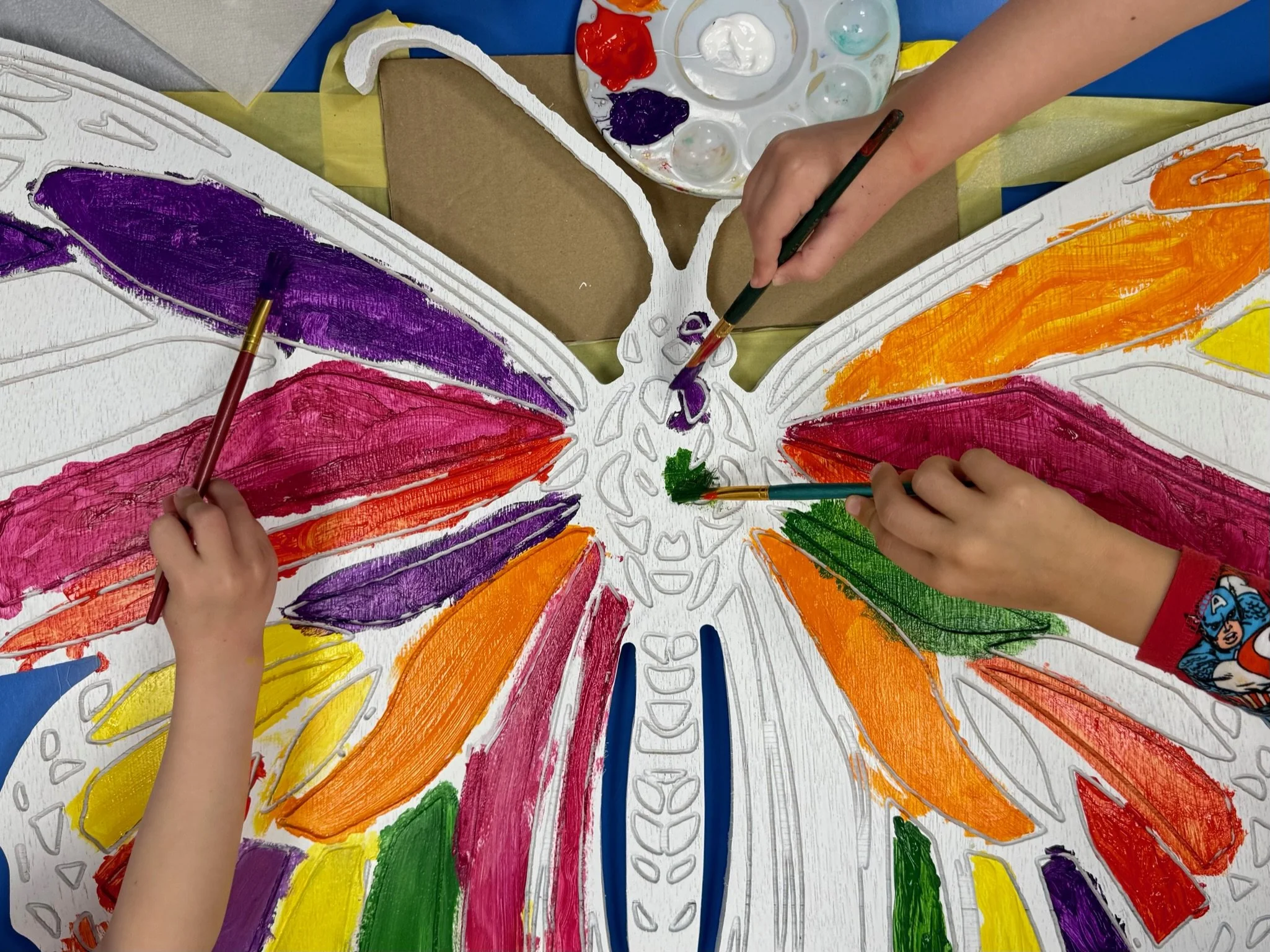 Children are painting a large butterfly on paper with colorful paints, including purple, red, orange, yellow, green, and pink.