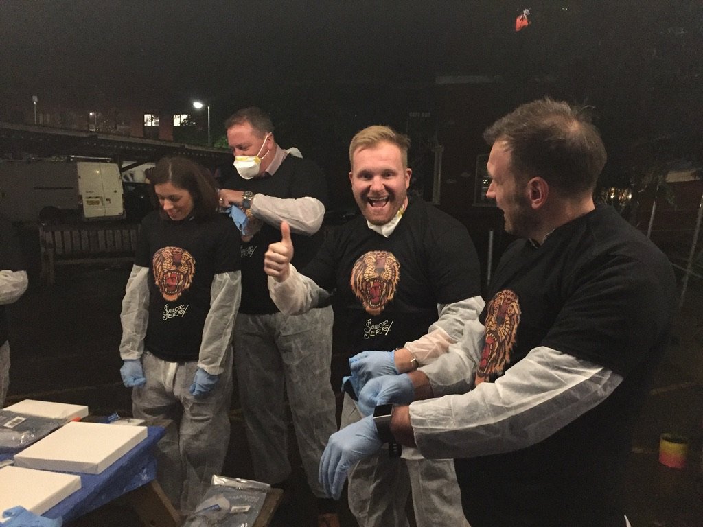 Four people inside at night, wearing black shirts with a lion graphic, protective gloves, and protective suits. One person in the middle is giving a thumbs-up and smiling at the camera. They appear to be preparing or handling items on a table.