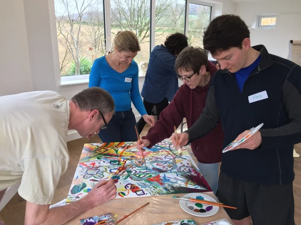 Group of five people collaborating on a large, colorful abstract painting inside a bright room with large windows and a view of trees outside.