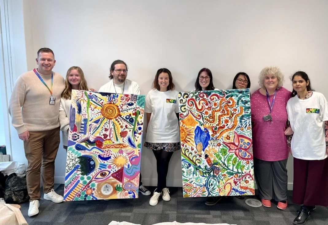 A group of eight people standing indoors, holding two large colorful abstract paintings with various swirling and geometric patterns.