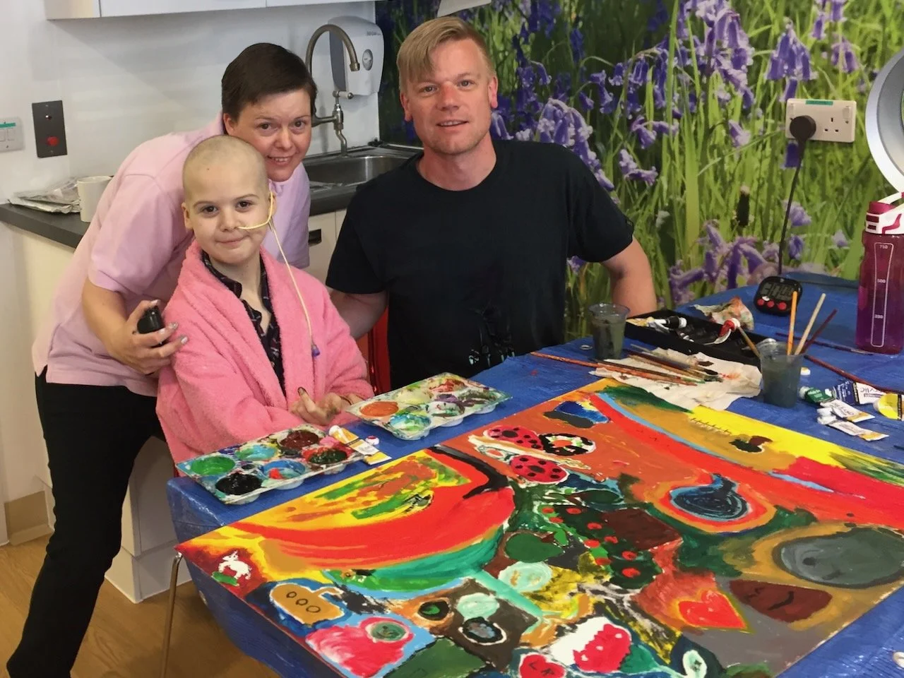 Three people in an art room with a colorful abstract painting on the table. A young girl with a nasal tube, wearing a pink robe, is sitting at the table. A woman in a pink shirt is standing behind her, and a man in a black shirt is sitting, all smiling.
