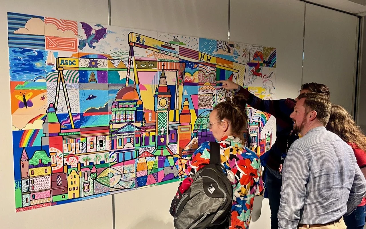 Group of people viewing and discussing a colorful, abstract mosaic mural of a cityscape with buildings and landmarks.