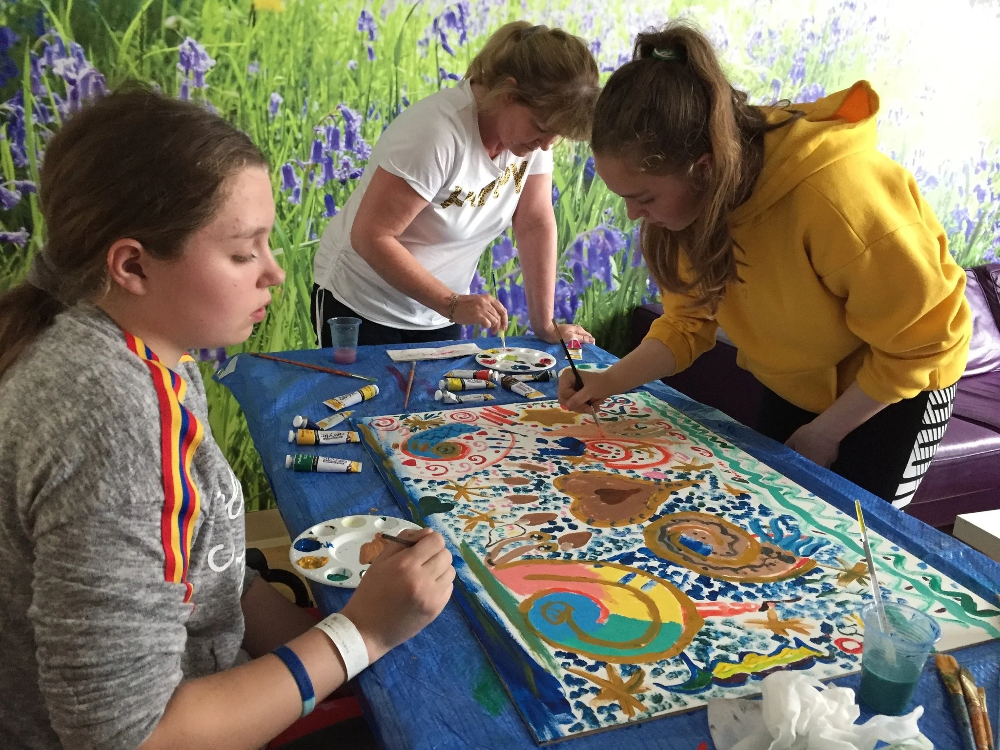 Group of young women and an older woman painting a large, colorful abstract artwork on a table at an indoor event with a nature-themed wall mural in the background.