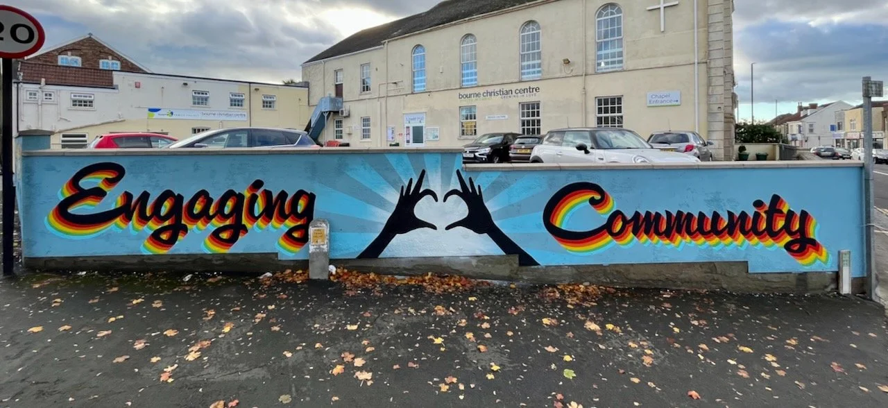 A mural on a wall with the words 'Engaging Community' in colorful, rainbow shadowed script, with two black hands forming a heart shape in the center, set against a blue sky background.