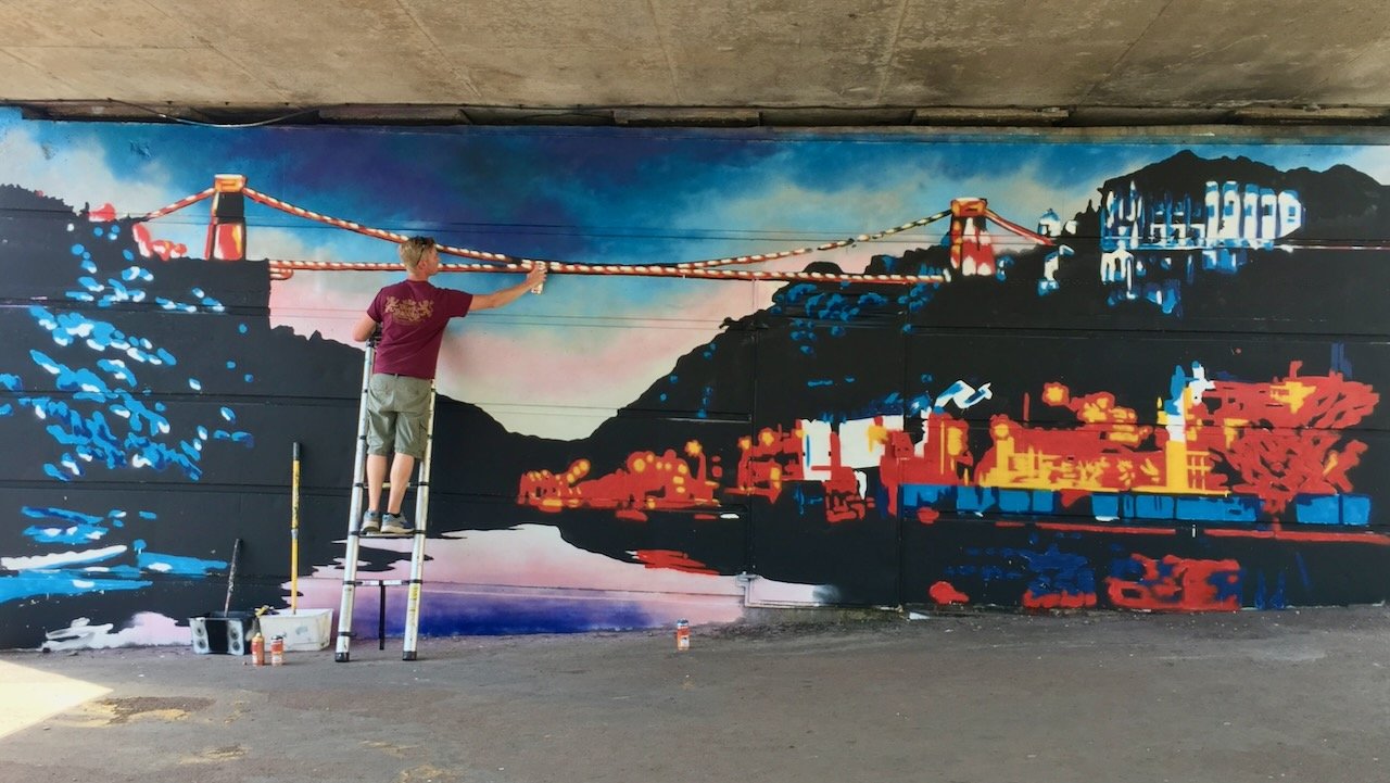 Artist painting a mural of the Golden Gate Bridge and city skyline under a sunset sky.