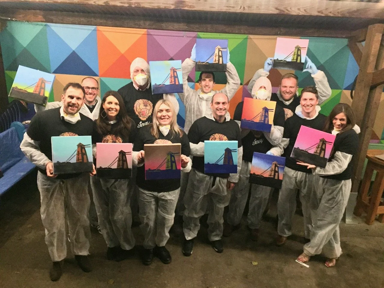 Group of people holding up paintings of the Golden Gate Bridge during a painting event, wearing protective smocks and face masks, in a colorful indoor setting.