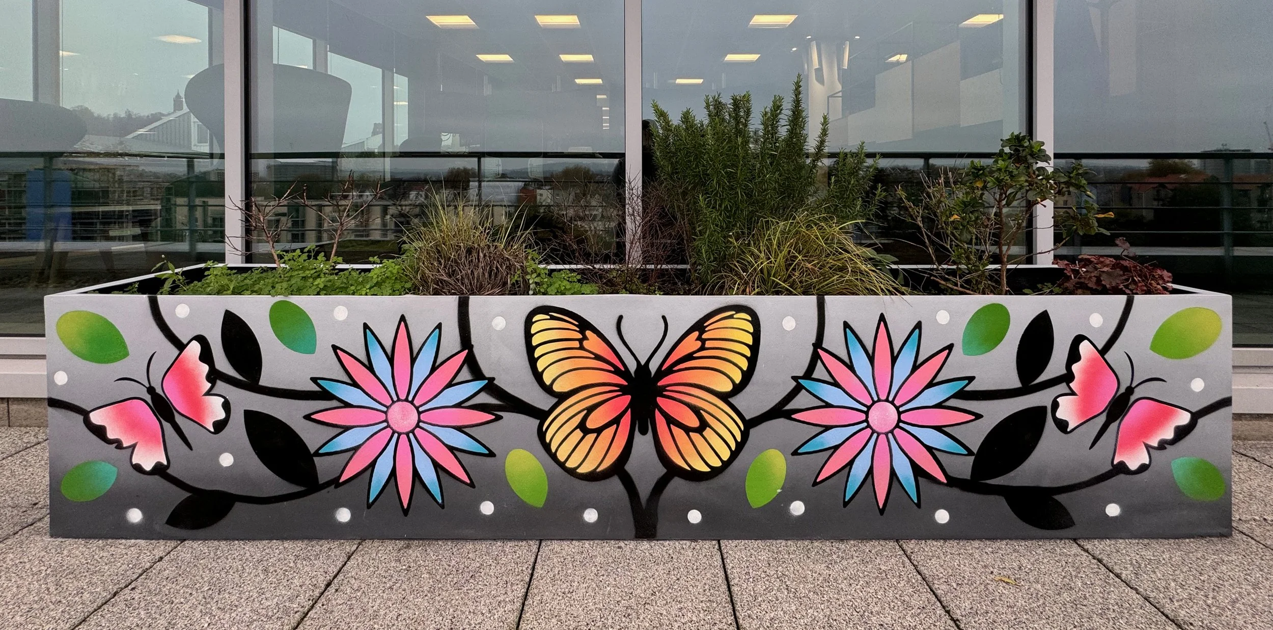 A decorative planter box with painted colorful flowers, butterflies, and green leaves on a grey background, containing various plants, placed outside a building with large glass windows.