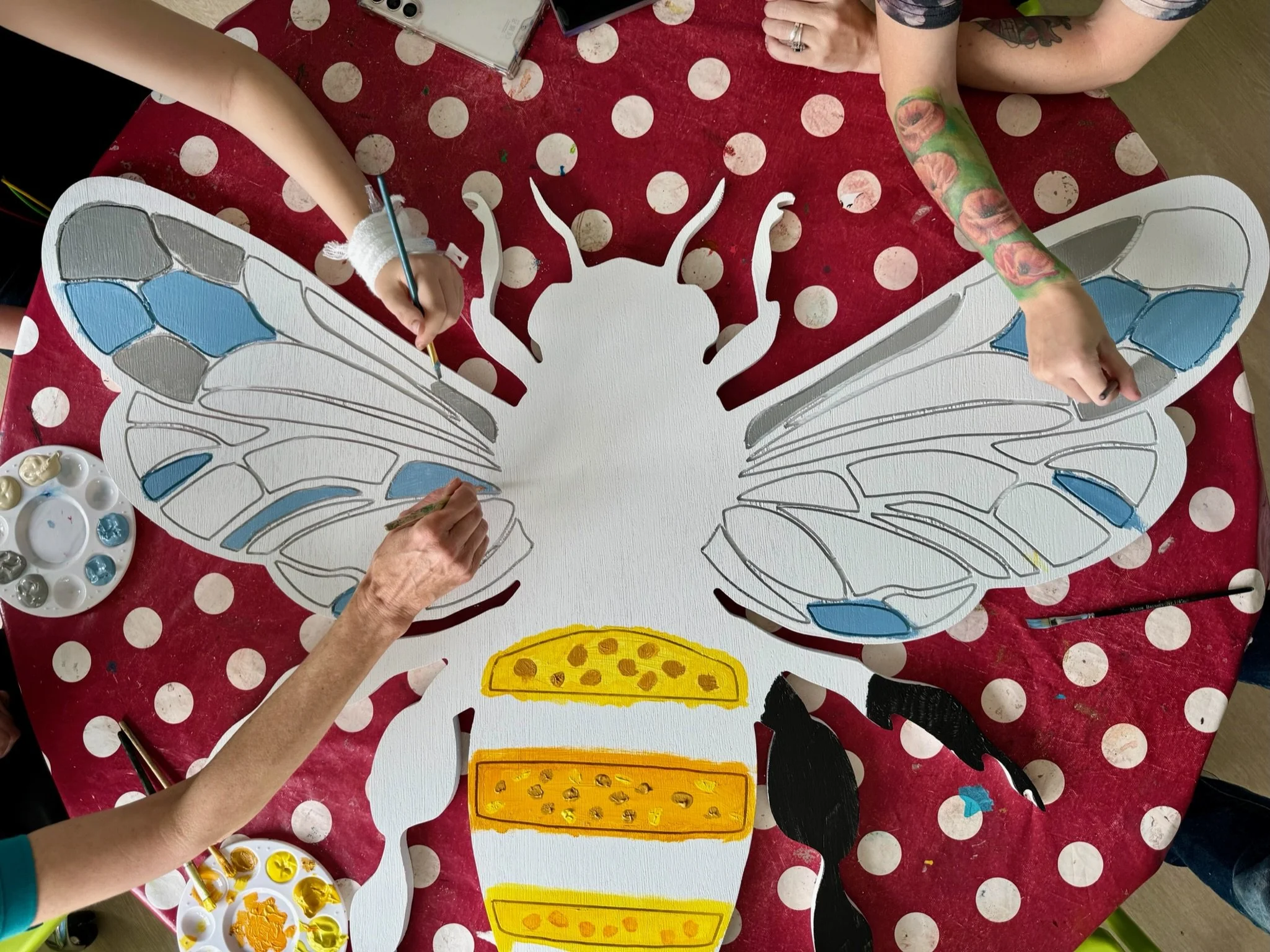 People painting a large butterfly cutout on a red polka dot tablecloth.