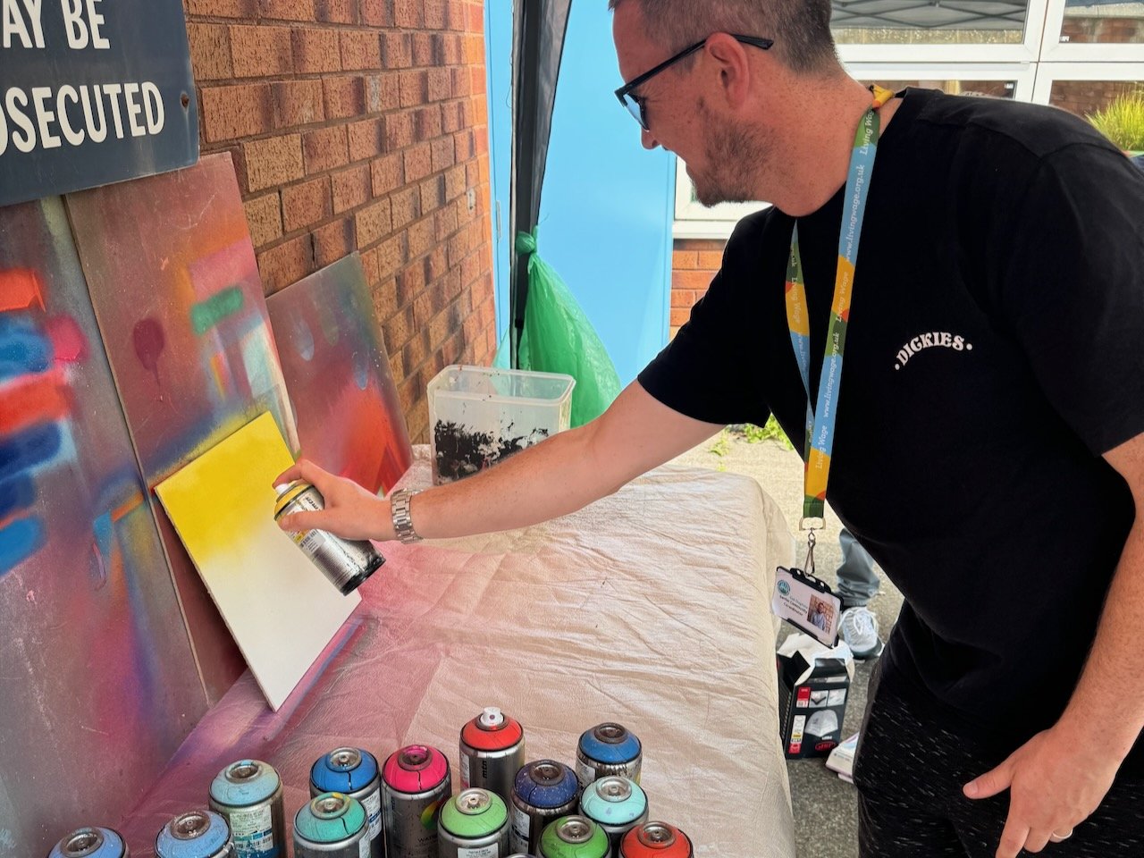 Man spray painting a canvas with yellow and red backgrounds, using spray paint cans on a table. He wears glasses, a black t-shirt, and a colorful lanyard.