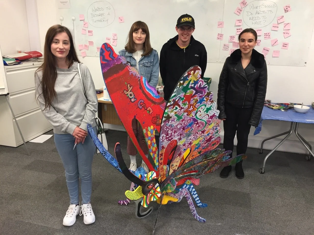 Group of four young people standing behind a large colorful butterfly art piece in an office or classroom. Two women on either side and two men in the middle, all smiling. Whiteboard with notes and pink sticky notes in the background.