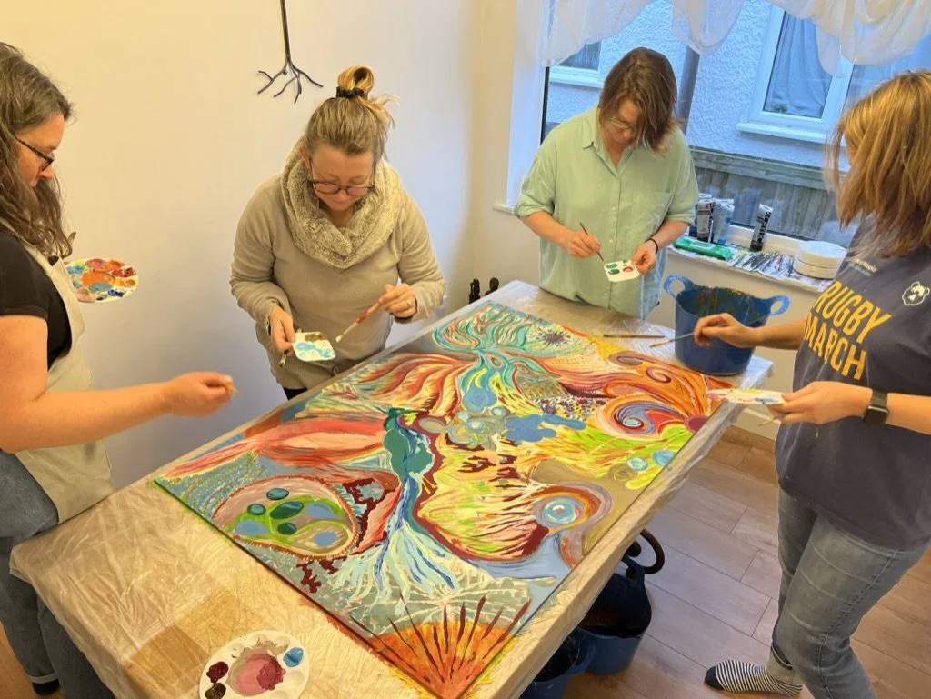 Four women are painting a colorful abstract artwork on a large canvas in a cozy indoor setting.