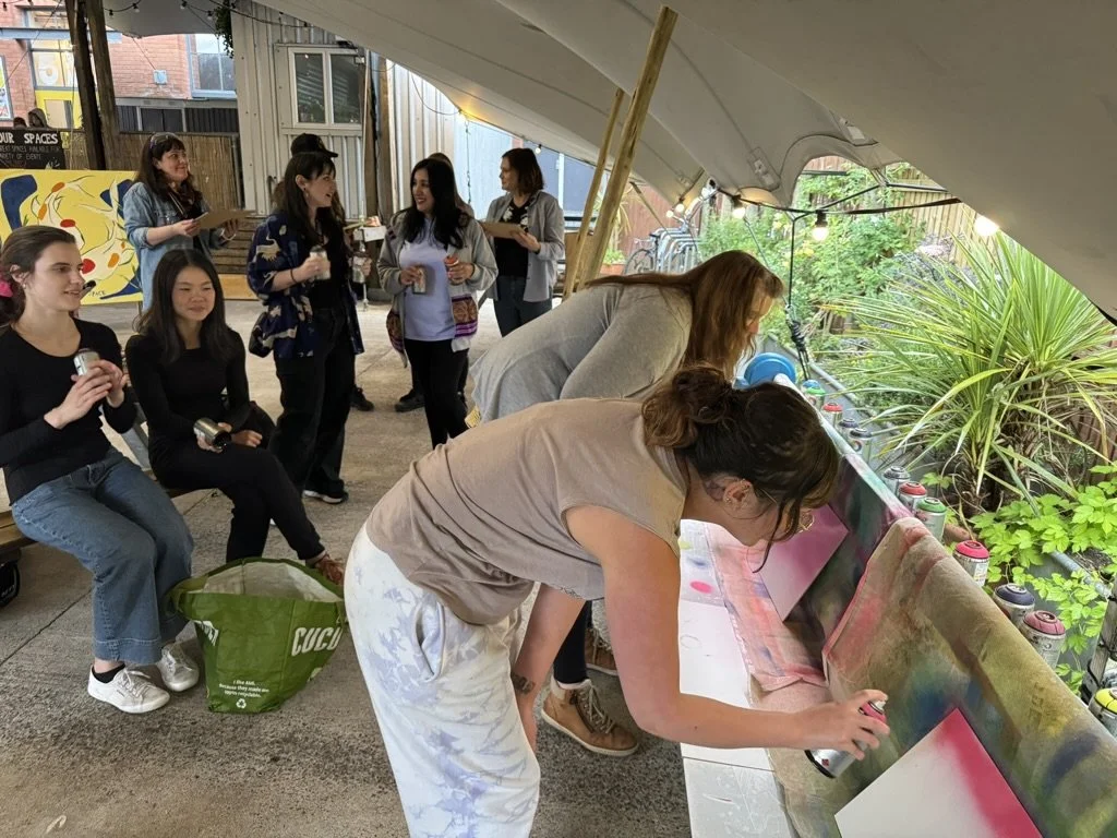 Group of women participating in a spray painting activity on a colorful canvas under a tent, with some women watching and others actively spray painting, surrounded by plants and outdoor string lights.