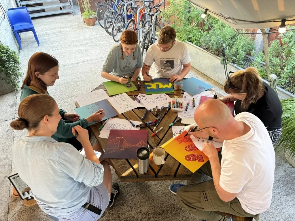 Six people sitting around a round wooden table outdoors, drawing and coloring posters, with various art supplies on the table, bicycles lined up in the background, and string lights overhead.