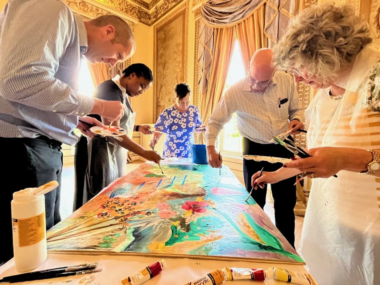 Group of people painting on a large canvas in an elegant room with gold accents and floral curtains.