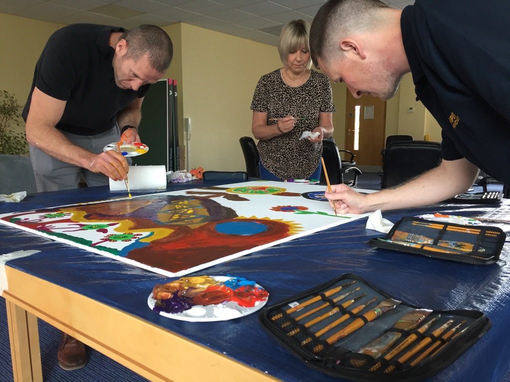 Three people, two men and one woman, are painting a large colorful mural on a table covered with plastic. The men are focused on their painting, while the woman looks on. Art supplies, including paintbrushes and paint palettes, are on the table.