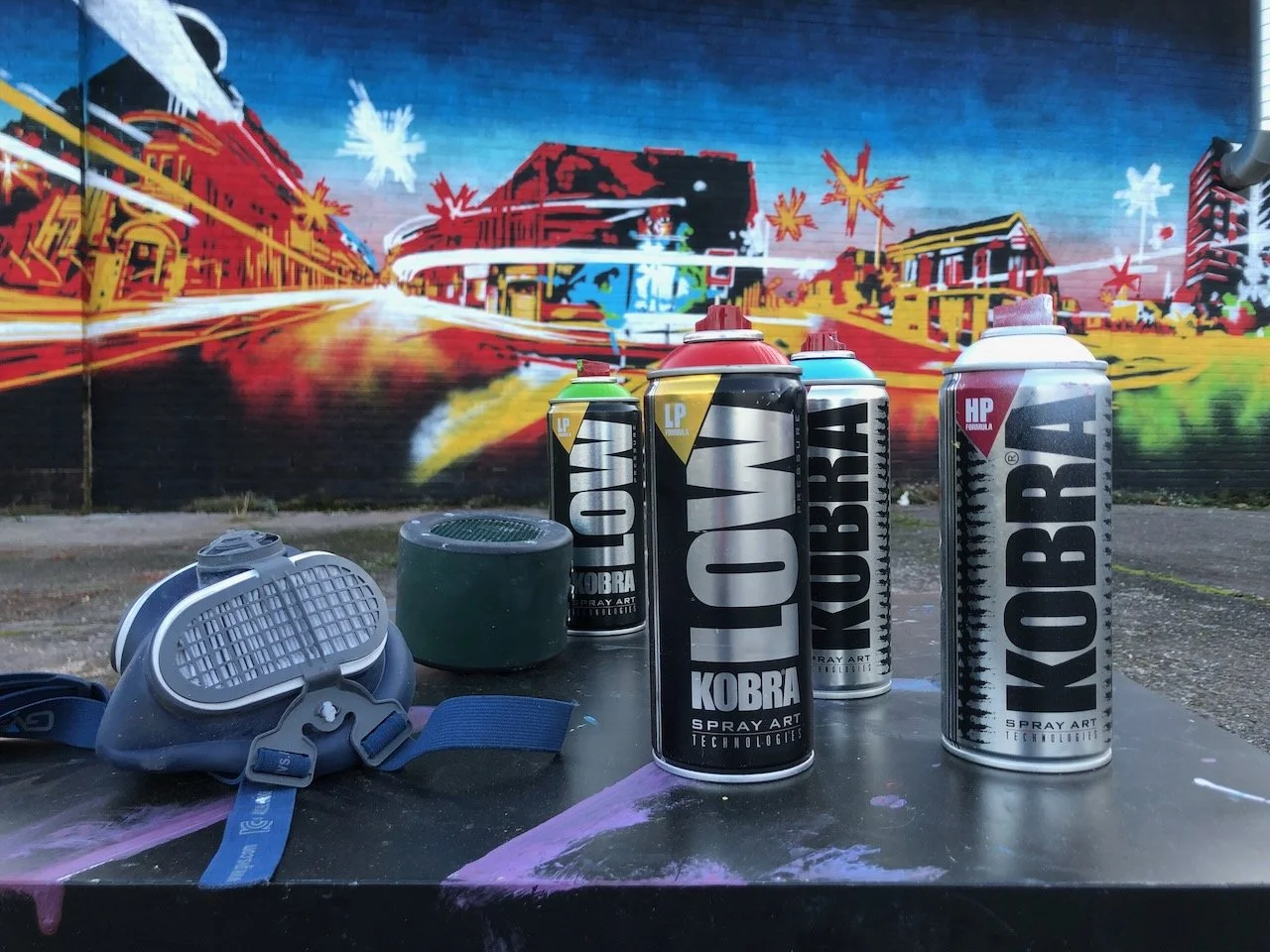 Four cans of Kobra spray paint placed on a table with various spray paint caps and a respirator mask nearby, against a colorful mural of a cityscape with buildings and star decorations.