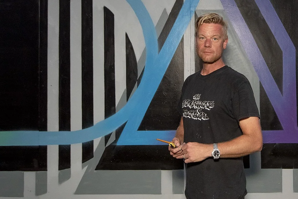 Man with blond hair and beard standing in front of a colorful abstract mural wall, holding a pencil and wearing a black T-shirt and a wristwatch.