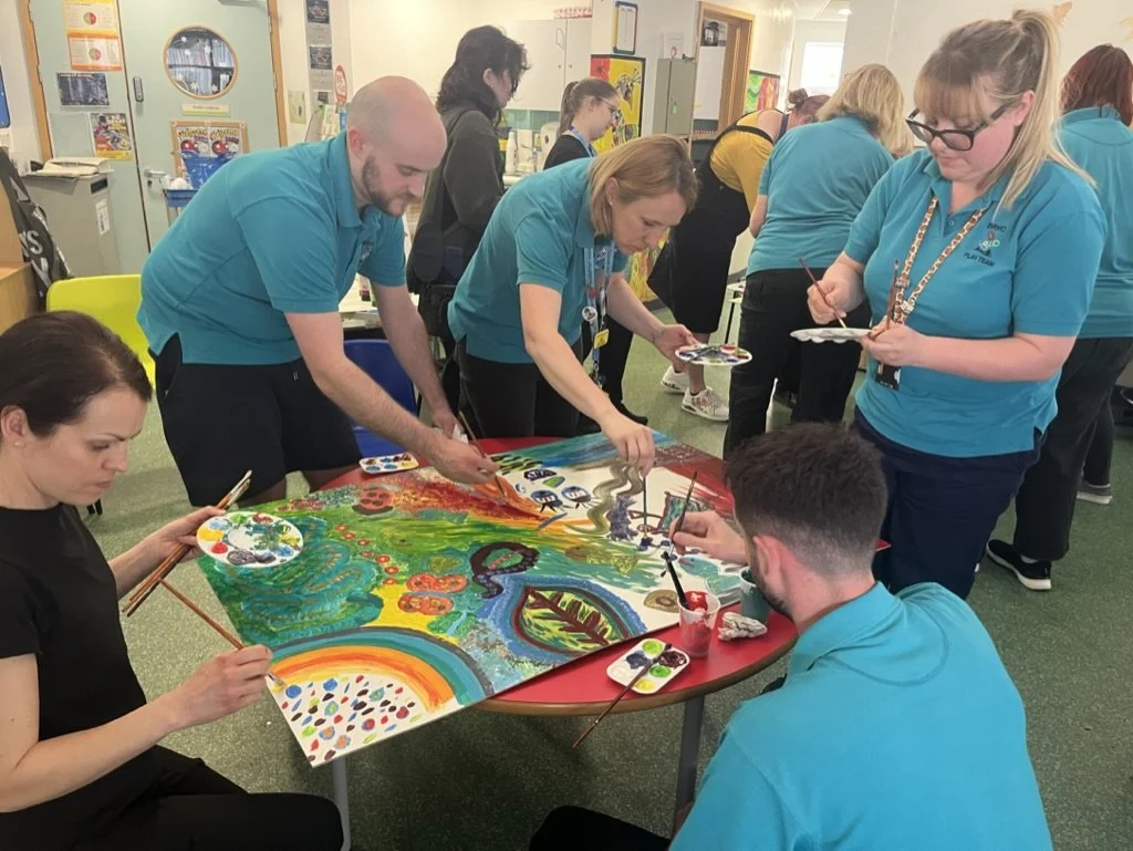 Group of people painting a colorful mural on a table in a classroom or art room.