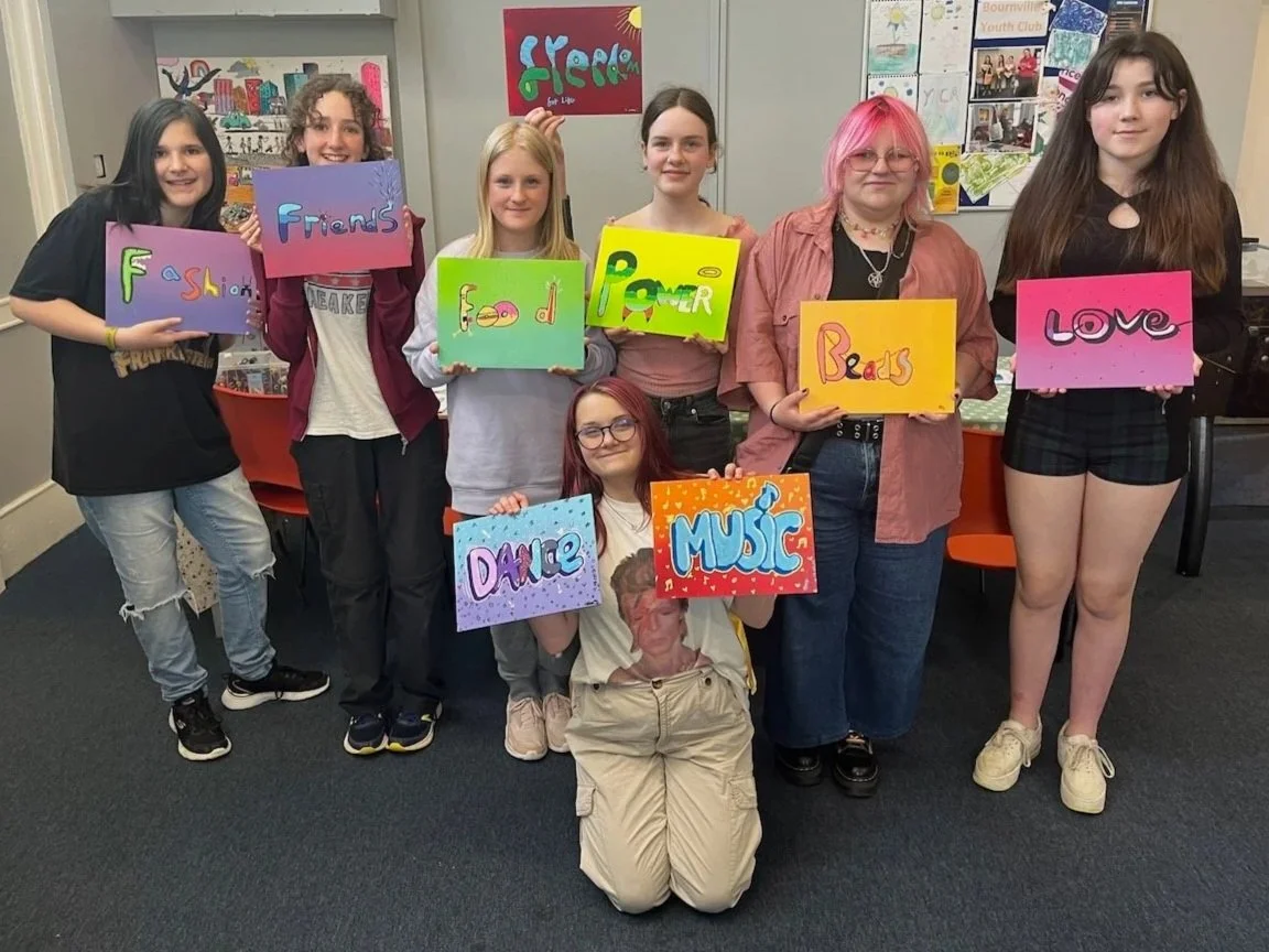 A group of eight young women standing indoors, holding colorful handmade signs with words like 'Fashion', 'Friends', 'Food', 'Power', 'Beads', 'Love', 'Dance', and 'Music'. They are smiling and appear to be in a creative or community center setting with artwork on the walls.