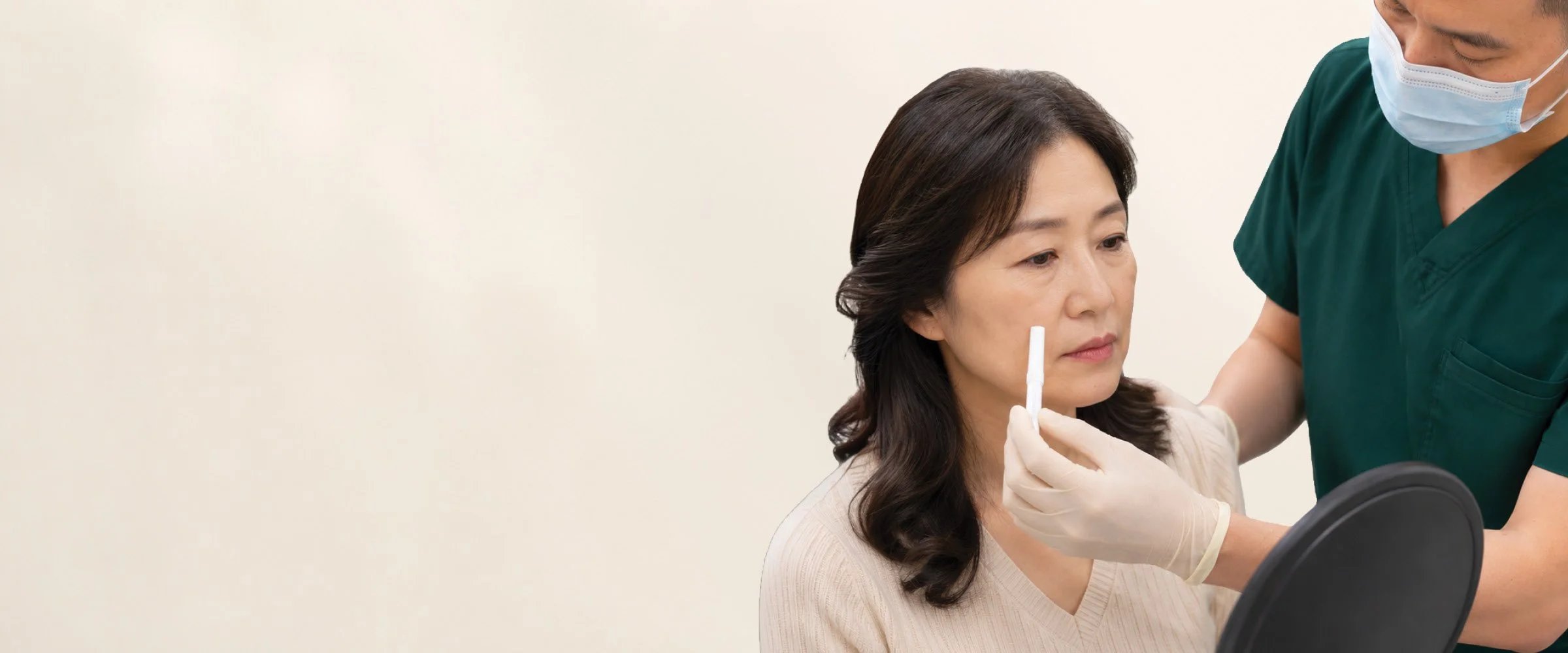 A healthcare professional in scrubs and a face mask is administering a nasal swab to a middle-aged woman with dark hair, seated and holding a mirror, against a plain light-colored background.