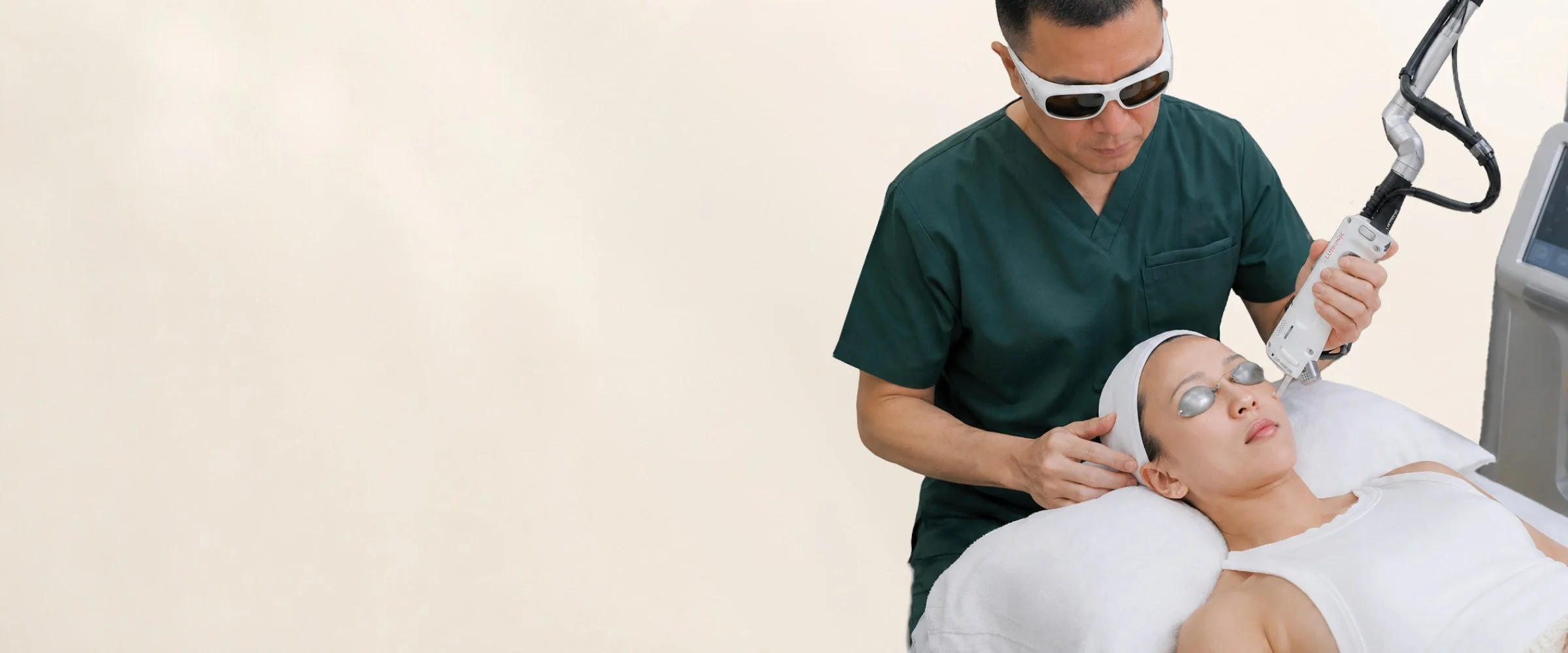 A medical professional in green scrubs and sunglasses performing a laser treatment on a woman lying on a treatment bed, with protective eye gear, in a clinical setting.