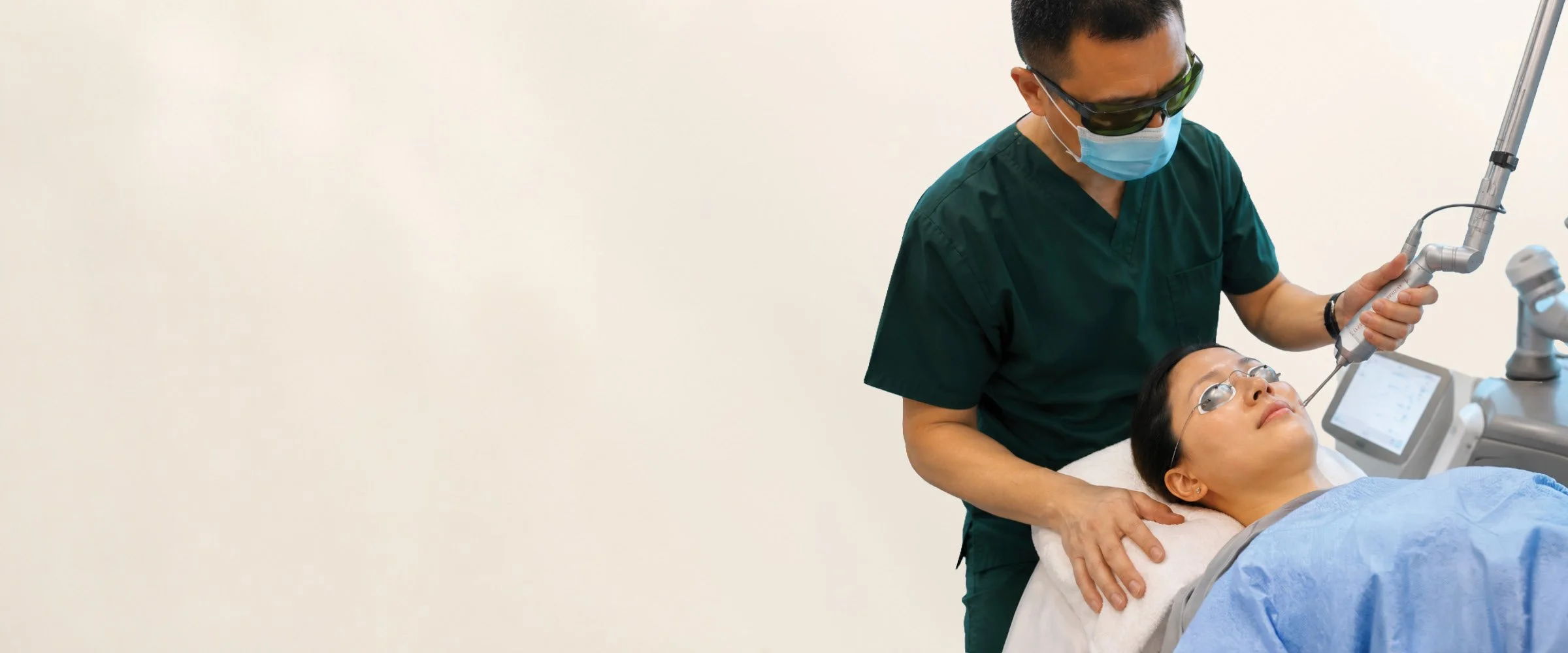 A healthcare professional in green scrubs, wearing a face mask and sunglasses, performs a procedure on a female patient lying on a medical bed with a medical device nearby. The patient is looking up, with glasses on and a blanket covering her.