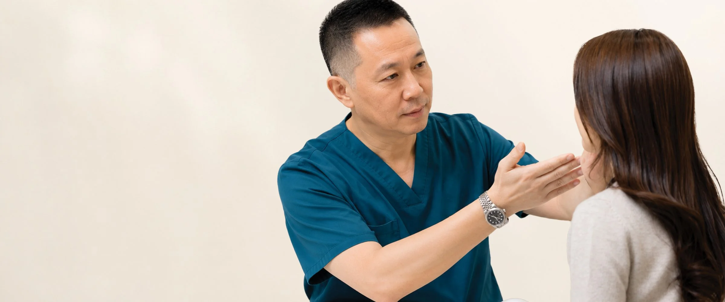 A male healthcare professional in blue scrubs examining a woman's neck in a clinical setting.
