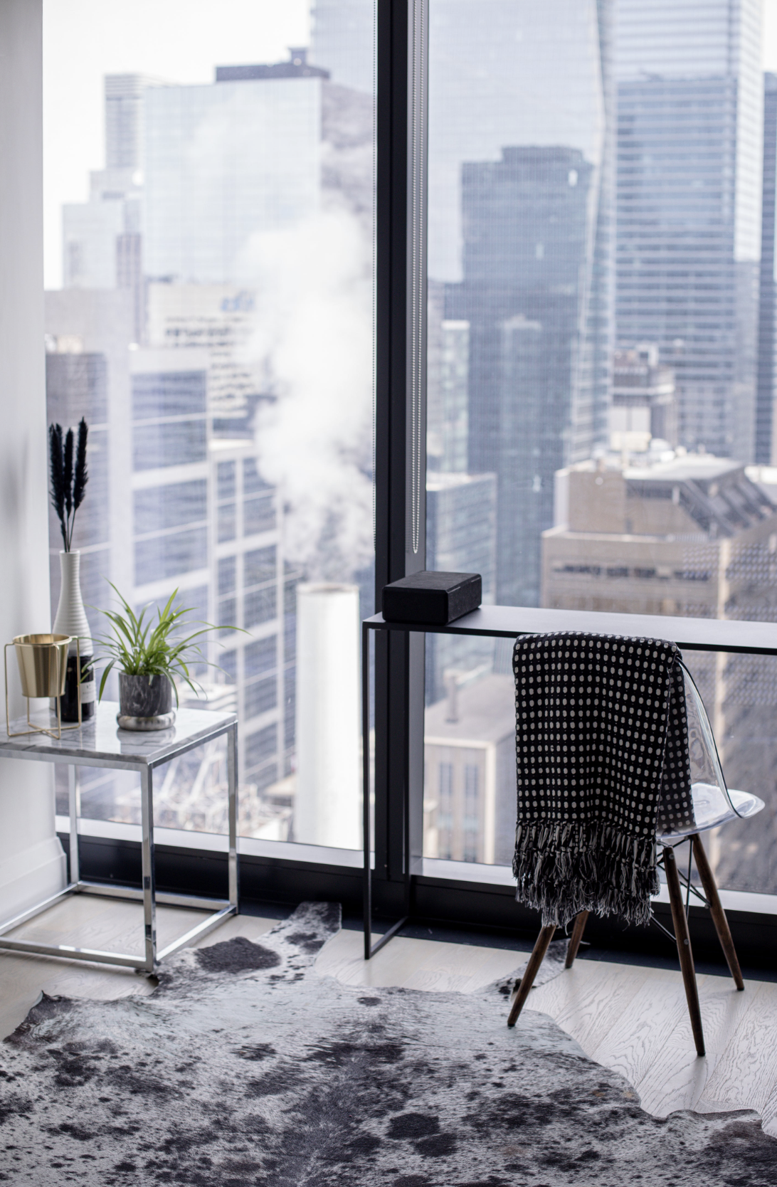 Modern office corner with a large cityscape view, a small table with potted plants, a checkerboard chair with a scarf, and a black speaker