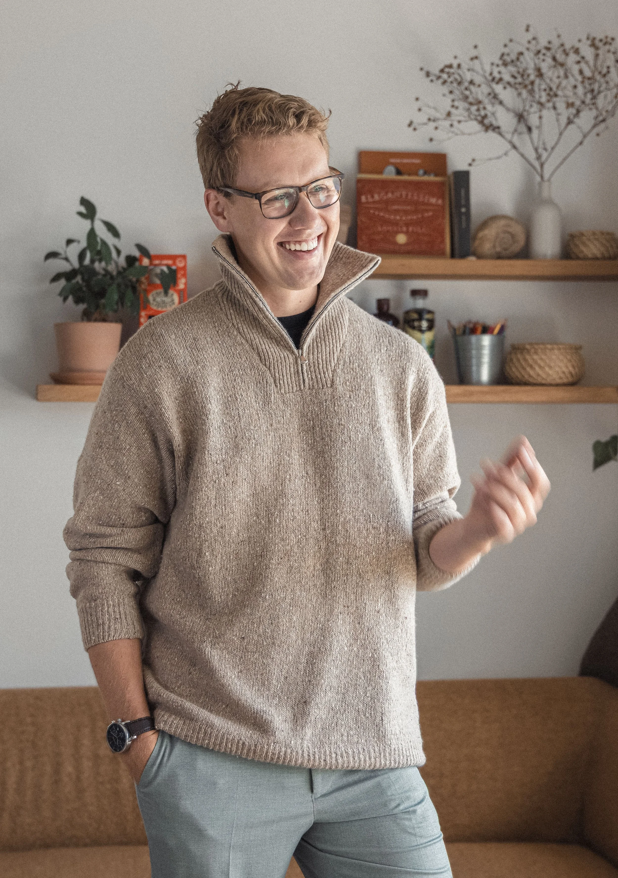 A smiling young man with glasses, wearing a beige zip-up sweater, standing indoors with a potted plant and shelves with books and decorative items in the background.
