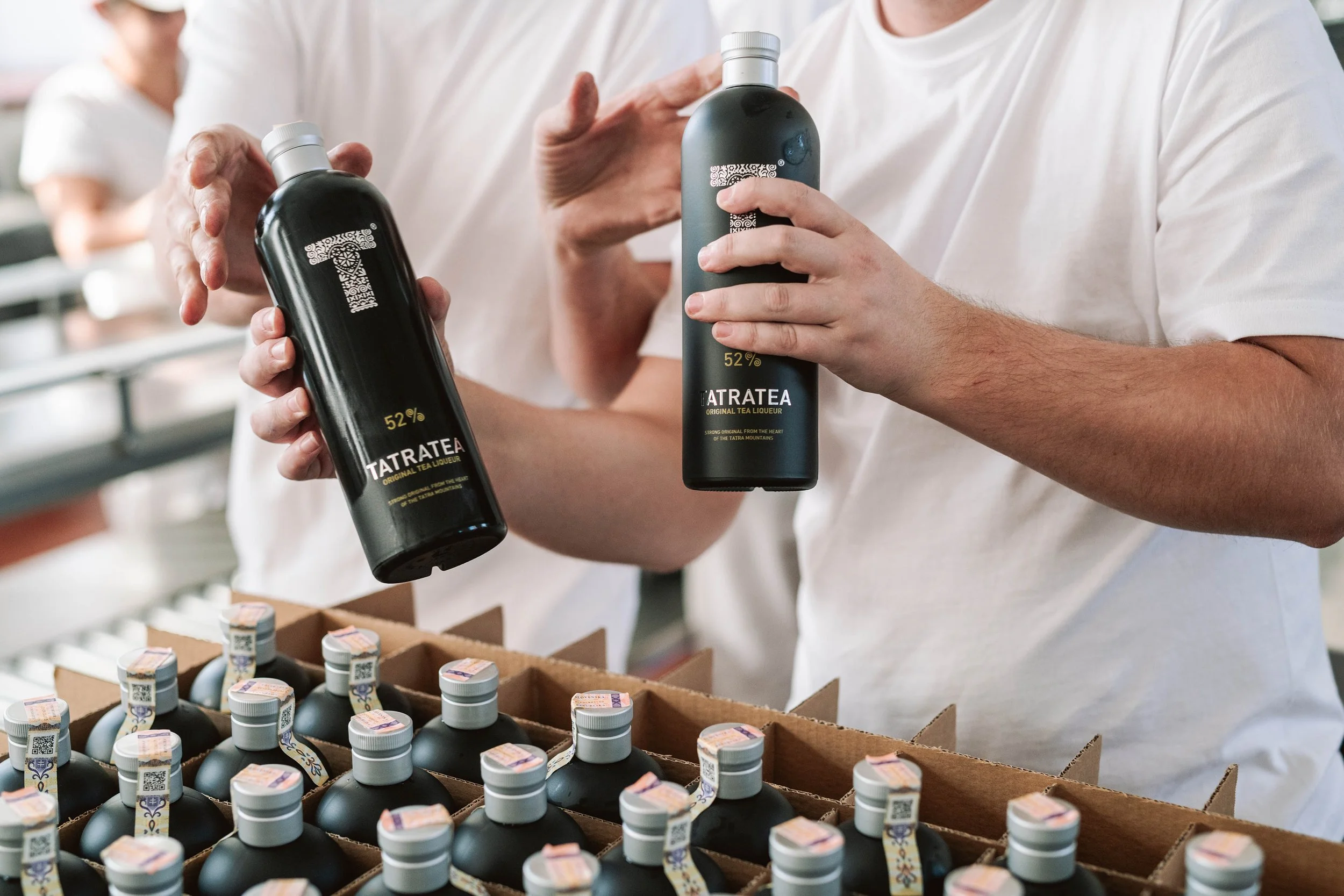 People holding bottles of Tatratea tea liqueur, in a production or packaging setting with boxes of bottles in the foreground.